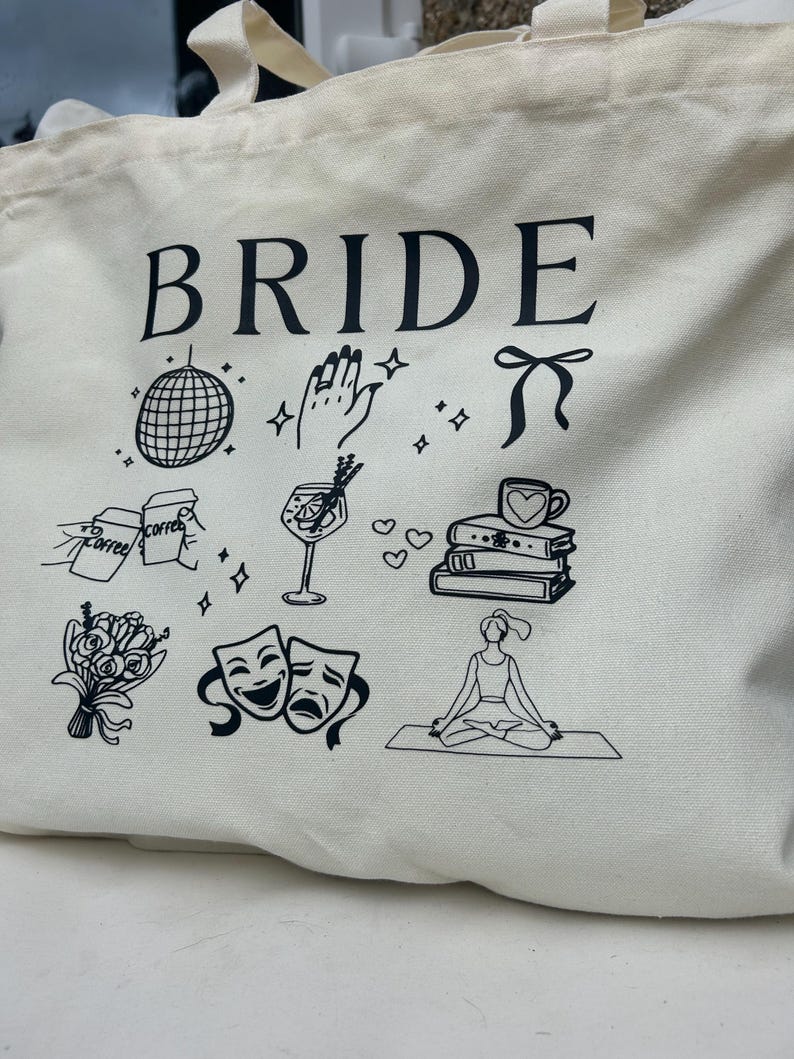 May include: Cream-colored tote bag with the word "BRIDE" in large black letters. The bag features black line drawings of a disco ball, a hand with a ring, a bow, coffee, a cocktail, books, flowers, theater masks, and a person meditating.
