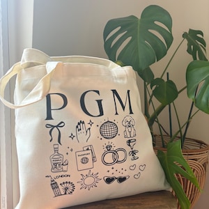 May include: Cream-colored tote bag with black illustrations and the letters "PGM". The bag features drawings of a bow, hand with sparkles, disco ball, dog, tequila bottle, passport, rings, wine glass, London eye, sun, and sunglasses.
