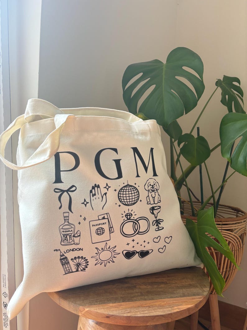 May include: Cream-colored tote bag with black illustrations and the letters "PGM". The bag features drawings of a bow, hand with sparkles, disco ball, dog, tequila bottle, passport, rings, wine glass, London eye, sun, and sunglasses.