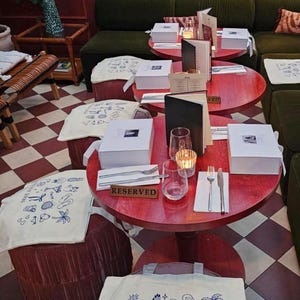 May include: A restaurant scene with small red tables, each set with glassware, cutlery, and a white box. White tote bags with blue illustrations and text reading "SICILY 2026" are visible.