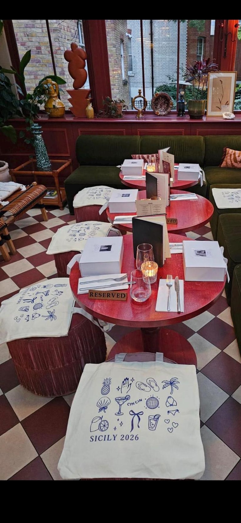 May include: A restaurant scene with small red tables, each set with glassware, cutlery, and a white box. White tote bags with blue illustrations and text reading "SICILY 2026" are visible.