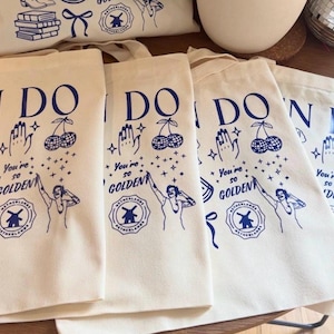 May include: Off-white canvas tote bags with blue illustrations and text. The design includes a dog, cherries, a hand, a watermelon slice, and the words "You're so GOLDEN." The bags have a Dutch windmill logo.
