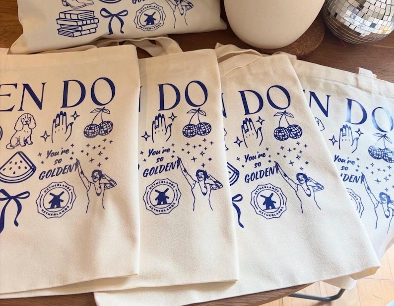 May include: Off-white canvas tote bags with blue illustrations and text. The design includes a dog, cherries, a hand, a watermelon slice, and the words "You're so GOLDEN." The bags have a Dutch windmill logo.