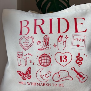 May include: Off-white tote bag with red graphics and text. The word "BRIDE" is at the top, with various illustrations below, including a heart, a cowboy boot, a disco ball, and a guitar. The text "MRS WHITMARSH TO BE" is at the bottom.
