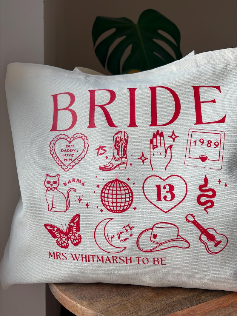 May include: Off-white tote bag with red graphics and text. The word "BRIDE" is at the top, with various illustrations below, including a heart, a cowboy boot, a disco ball, and a guitar. The text "MRS WHITMARSH TO BE" is at the bottom.