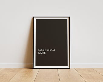 Könnte beinhalten: Ein gerahmtes schwarzes Poster mit dem Text "LESS REVEALS MORE." Das Poster hat einen weißen Rand und einen schwarzen Rahmen. Es lehnt an einer weißen Wand auf einem Holzboden.