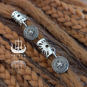 Könnte beinhalten: Silberfarbene Haaraccessoires aus Metall mit aufwendigen kreisförmigen Designs. Sie sind an einem braunen Zopf befestigt. Das Bild enthält das Logo "Falkya" mit einer Wikingerschiff-Illustration.