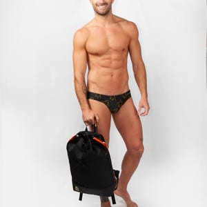 May include: A black backpack with orange detailing is held by a person wearing black swim briefs with a gold pattern. The person is standing in front of a white backdrop.