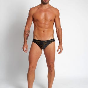Peut inclure: Un slip de bain noir avec un motif doré. Le maillot de bain est porté par une personne avec un tatouage sur le bras. Le fond est blanc uni.