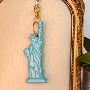 May include: A Statue of Liberty keychain in a light blue and white color scheme. The keychain is attached to a gold-colored clasp and ring. The background is a gold-framed, cream-colored surface.