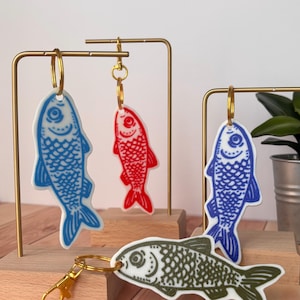 May include: Four fish-shaped keychains made of white material with printed designs. One keychain features a light blue fish, one a red fish, one a dark blue fish, and one an olive green fish. Each keychain includes a gold-toned metal ring and clasp.