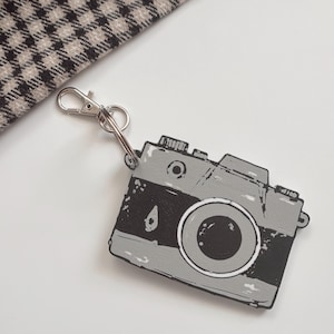 May include: A gray and black camera-shaped keychain with a silver clasp. The keychain has a vintage design, with a lens and details of a classic camera. A patterned fabric is visible in the background.