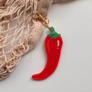 May include: A red chili pepper charm with a green stem and gold-colored clasp attached to a beige crocheted bag. The charm is a vibrant red color and has a glossy finish. The bag has a net-like design.