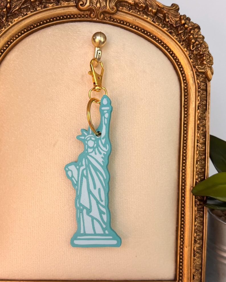 May include: A Statue of Liberty keychain in a light blue and white color scheme. The keychain is attached to a gold-colored clasp and ring. The background is a gold-framed, cream-colored surface.