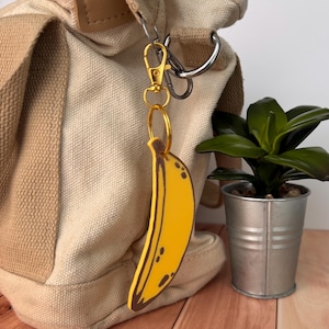 May include: A yellow banana-shaped keychain with brown details hangs from a gold-colored clasp attached to a beige canvas bag. The keychain is next to a small potted plant in a silver bucket.