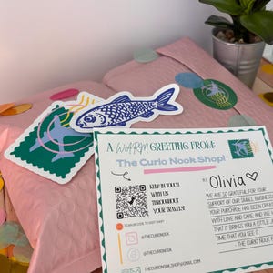 May include: A pink bubble mailer with stickers and a greeting card. The stickers include a blue fish and a green stamp design. The card reads "A Warm Greeting From: The Curio Nook Shop!" and includes social media handles.