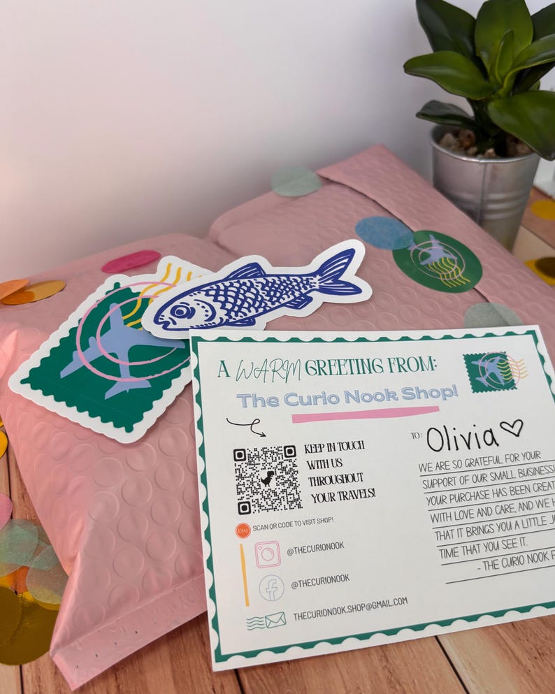 May include: A pink bubble mailer with stickers and a greeting card. The stickers include a blue fish and a green stamp design. The card reads "A Warm Greeting From: The Curio Nook Shop!" and includes social media handles.