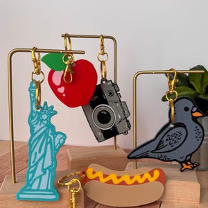 May include: A collection of novelty keychains featuring iconic symbols. Includes a light blue Statue of Liberty, a red apple, a gray camera, a gray bird, and a hot dog with mustard. Each keychain has a gold-colored clasp.