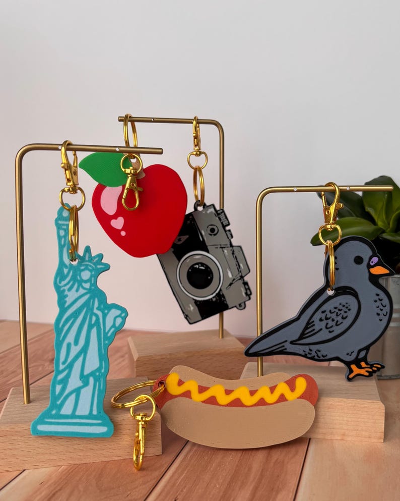 May include: A collection of novelty keychains featuring iconic symbols. Includes a light blue Statue of Liberty, a red apple, a gray camera, a gray bird, and a hot dog with mustard. Each keychain has a gold-colored clasp.