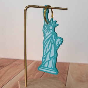 May include: A turquoise Statue of Liberty keychain with white detailing hangs from a gold-colored metal stand. The keychain is attached to a gold-colored ring. The stand is mounted on a wooden base.
