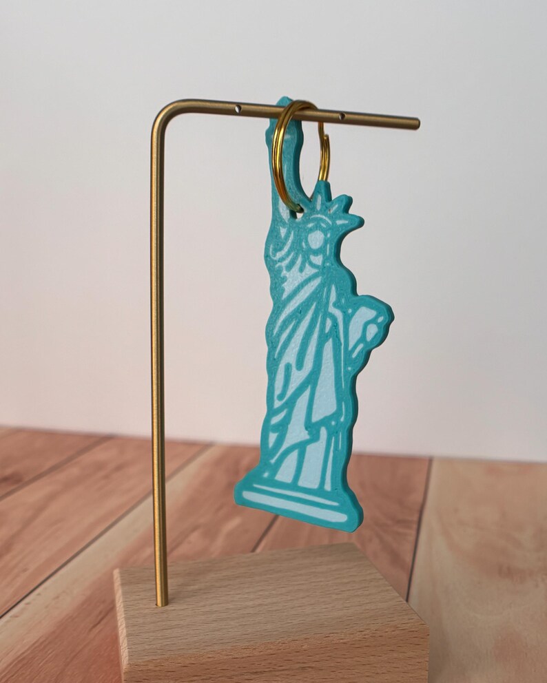 May include: A turquoise Statue of Liberty keychain with white detailing hangs from a gold-colored metal stand. The keychain is attached to a gold-colored ring. The stand is mounted on a wooden base.