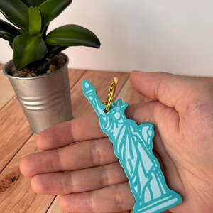 May include: A turquoise Statue of Liberty keychain with a gold-colored ring. The keychain is held in a hand, with a small potted plant in the background. The keychain is a souvenir.