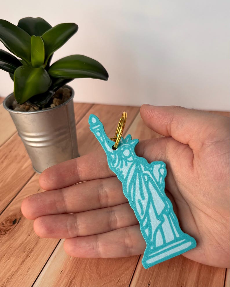 May include: A turquoise Statue of Liberty keychain with a gold-colored ring. The keychain is held in a hand, with a small potted plant in the background. The keychain is a souvenir.