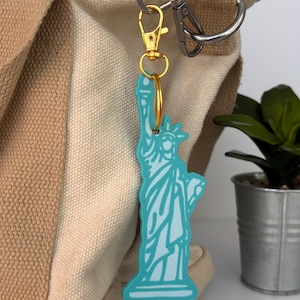 May include: A light blue Statue of Liberty keychain with a gold-colored clasp and ring. The keychain is attached to a beige and tan tote bag. A small potted plant is in the background.