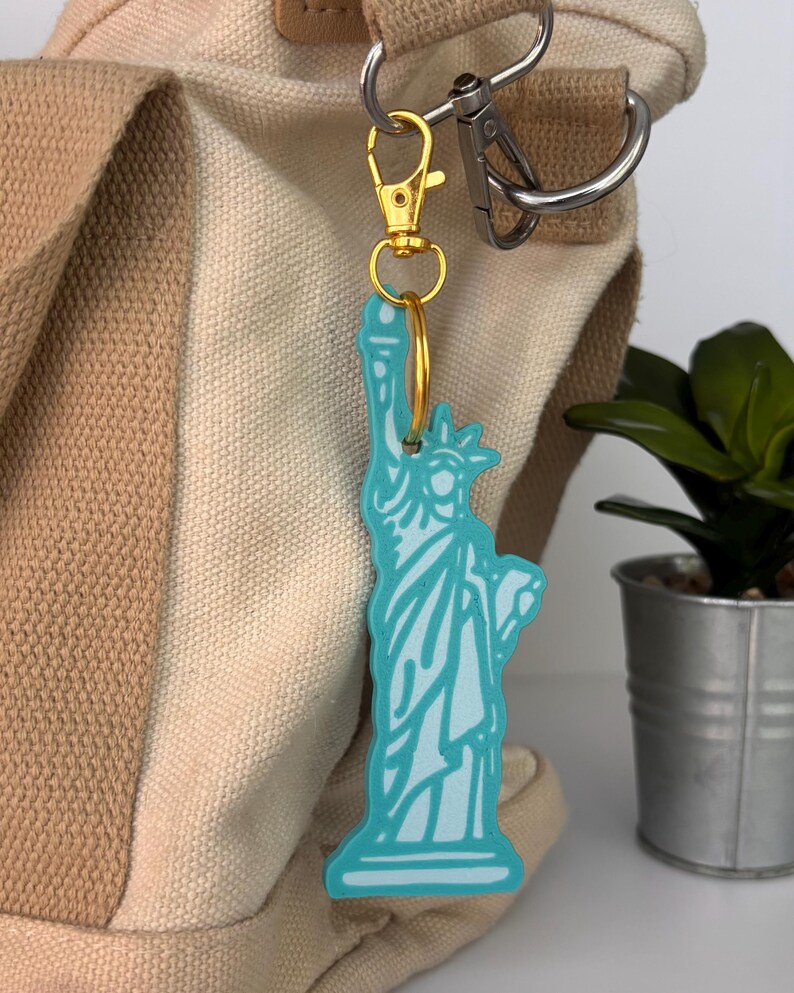 May include: A light blue Statue of Liberty keychain with a gold-colored clasp and ring. The keychain is attached to a beige and tan tote bag. A small potted plant is in the background.