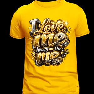May include: A yellow t-shirt with the phrase "I Love Me heavy on the me" in a metallic, 3D design. The text features silver, gold, and black accents with starburst details, creating a bold and eye-catching graphic.
