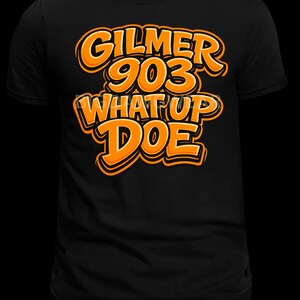 Puede incluir: Camiseta negra con texto naranja "GILMER 903 WHAT UP DOE". El texto tiene un diseño audaz y contorneado. La camiseta es de estilo cuello redondo.