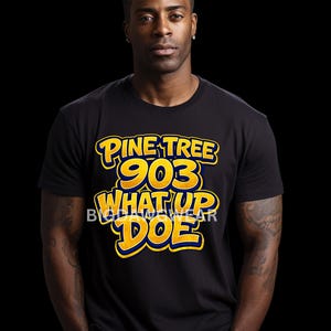 May include: Black t-shirt featuring the text "PINE TREE 903 WHAT UP DOE" in yellow and blue. The shirt is worn by a person with dark hair and a watch.