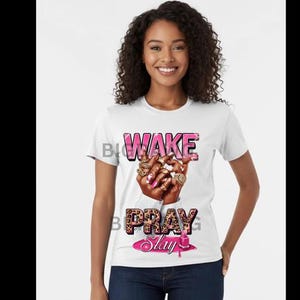 Puede incluir: Camiseta blanca con las palabras "WAKE PRAY SLAY" en letras rosas y marrones. El gráfico incluye una mano adornada con anillos y esmalte de uñas rosa. La modelo lleva vaqueros azules.