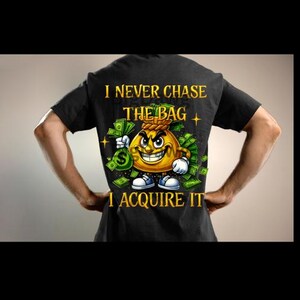 Pode incluir: Camiseta preta com um personagem de saco de dinheiro de desenho animado sorrindo, segurando um saco de dinheiro e cercado por dinheiro. O texto "I NEVER CHASE THE BAG I ACQUIRE IT" é impresso em ouro.