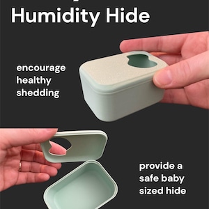 Puede incluir: Un recipiente rectangular verde claro con una abertura en forma de corazón en la tapa. El texto "Baby Humidity Hide" está en la parte superior. El texto adicional incluye "encourage healthy shedding", "provide a safe baby sized hide" y "Smooth corners for easy cleaning".
