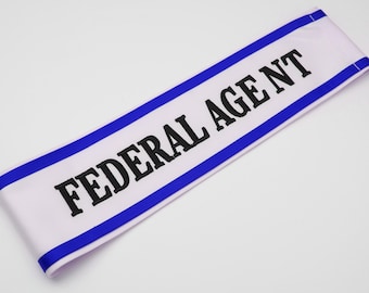 May include: White sash with the words "FEDERAL AGENT" in bold black letters. The edges are trimmed with a thin blue border. The sash is made of a smooth, likely satin-like material.
