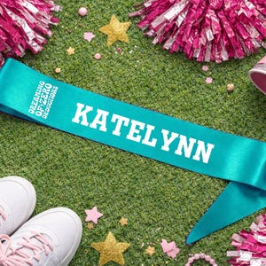 May include: A teal satin sash with the name "KATELYNN" in bold white letters. The sash also features the text "DREAMING OF ZERO DEDUCTIONS". The sash is surrounded by cheerleading accessories, including pom-poms, a megaphone, and sneakers, on a green surface.