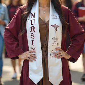 May include: A maroon graduation gown with a white sash that reads "NURSING" vertically. The sash also displays a medical symbol, the text "Class of 2025", and the words "Cum Laude". The gown is worn over a brown dress.