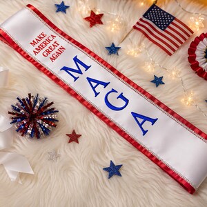 May include: A white satin sash with red trim and the embroidered text "MAKE AMERICA GREAT AGAIN" and "MAGA" in blue. The image includes an American flag, star-shaped decorations, and a red, white, and blue rosette.