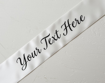 May include: White satin sash with the text "Your Text Here" in a black, elegant script. The sash is angled across a textured, off-white surface. The text is centered on the sash.