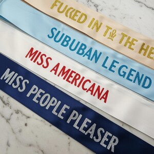 May include: An assortment of satin sashes in various colors, each emblazoned with glitter text. The sashes include phrases like "FUCKED IN THE HEAD," "SUBURBAN LEGEND," "MISS AMERICANA," and "MISS PEOPLE PLEASER." The sashes are arranged diagonally against a marble background.