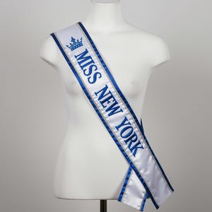 May include: A white satin sash with blue trim and text that reads "MISS NEW YORK" in blue. A blue crown graphic is above the text. The sash is draped over a white mannequin.