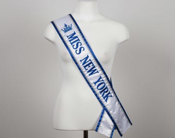 May include: A white satin sash with blue trim and text that reads "MISS NEW YORK" in blue. A blue crown graphic is above the text. The sash is draped over a white mannequin.