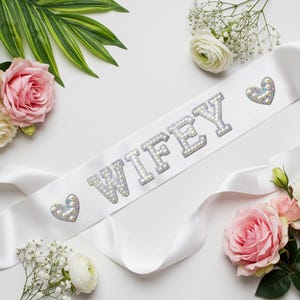 May include: A white satin sash with the word "WIFEY" spelled out in sparkling, rhinestone-like letters. Two heart-shaped embellishments with iridescent accents flank the text. The sash is surrounded by pink and white roses, greenery, and small white flowers.