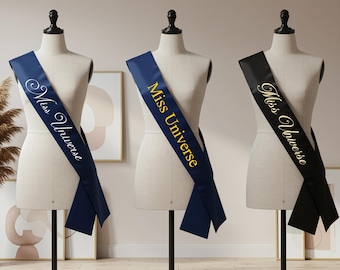 May include: Three satin sashes draped over mannequins. One is navy blue with white text, another is navy blue with gold text, and the third is black with white text. All sashes read "Miss Universe" in elegant script.