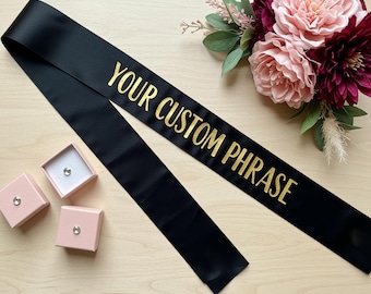 May include: Black satin sash with gold text that reads "YOUR CUSTOM PHRASE." The sash is displayed with three small pink boxes, each containing a small, round, silver-colored gem. A bouquet of pink and burgundy flowers is in the background.