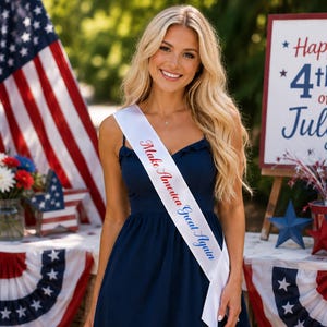 May include: A woman wearing a navy blue dress and a white sash with the text "Make America Great Again." The background includes an American flag, a sign that says "Happy 4th of July!", and patriotic decorations.