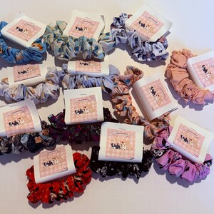 May include: A collection of colorful scrunchies in various patterns, including floral, polka dot, and abstract designs. Each scrunchie is paired with a small, square card featuring a cartoon illustration. The scrunchies are made of fabric and are designed for hair styling.