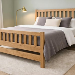 May include: A wooden bed frame with a slatted headboard and footboard. The bed is covered with a white comforter and a gray quilt. There are gray and cream-colored pillows. The bed sits on a light-colored rug.