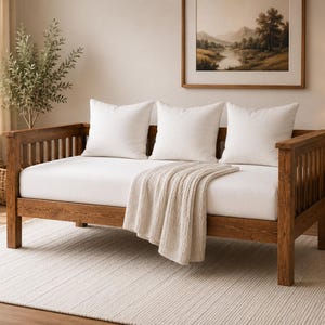 Peut inclure: Un lit de repos en bois avec un cadre à lattes et une finition marron chaud. Le lit de repos comprend un matelas blanc épais, trois coussins carrés blancs disposés le long du dossier et un plaid beige texturé drapé sur le bord avant.